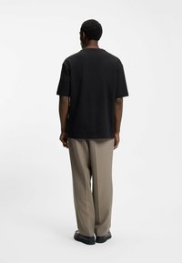Uomo in camicia nera a maniche corte, pantaloni beige e scarpe nere in piedi di spalle su uno sfondo bianco uniforme.