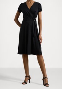 Zwarte wikkeljurk van zachte stof met korte mouwen, V-hals en een strik in de taille. Gaat gepaard met zwarte hoge hakken.