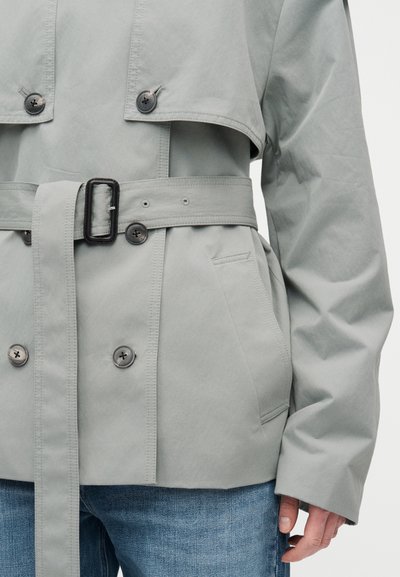 Ανοιχτό γκρι καμπαρντίνα με ζώνη και διπλή σειρά κουμπιών, πλαϊνές τσέπες, φορεμένο με μπλε τζιν και το δεξί χέρι χαλαρά στο πλάι.