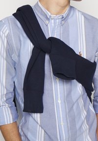 Marinblå sweatshirt som hänger över en ljusblå och vit randig skjorta med krage. Skjortan har en liten logotyp på bröstet.