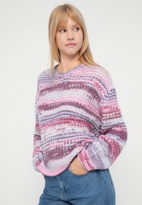 Woman wearing a multicolored striped knit sweater with hands in pockets, looking to the side against a plain light background.