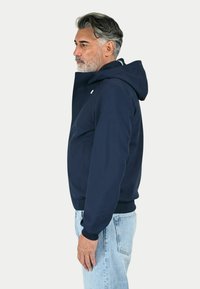 Giacca con cappuccio blu navy in tessuto liscio con zip frontale, polsini a costine e un piccolo logo sul petto. Abbinata a jeans azzurri.