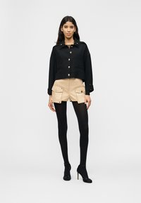 Γυναίκα που φοράει μαύρο κοντό σακάκι, μπεζ σορτς καρό, μαύρο καλσόν και μαύρα σανδάλια με τακούνια, σε απλό φωτεινό φόντο.