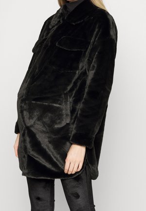 Persoon die een zwarte oversized faux fur jas draagt met borstzakken en zwarte leggings tegen een effen achtergrond.