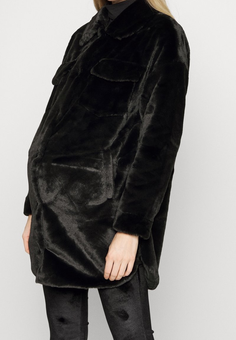 Personne portant une veste en fausse fourrure noire oversize avec des poches sur la poitrine et des leggings noirs sur un fond uni.