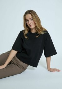 Black short-sleeve top made of cotton with a relaxed fit and round neckline, paired with brown trousers, set against a light background.