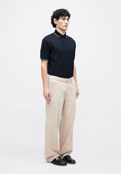 Μαύρο πόλο μπλουζάκι με λευκό γιακά και μανσέτες, συνδυασμένο με ανοιχτό μπεζ παντελόνι φαρδιάς γραμμής και μαύρα παπούτσια, σε ουδέτερο φόντο.