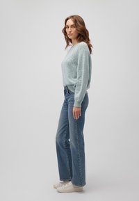 Hellblauer, gestrickter Pullover mit lockerer Passform und Spitzenmuster an den Ärmeln, kombiniert mit hochgeschnittenen blauen Jeans und weißen Sneakers.