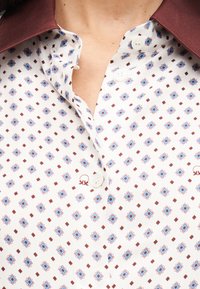 Άσπρο πουκάμισο με κουμπιά, με γεωμετρικό σχέδιο σε μπλε και καφέ αποχρώσεις, ανοιχτός γιακάς και ορατό σκούρο βυσσινί ύφασμα εσωτερικού γιακά.