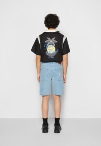 Czarny t-shirt z krótkim rękawem z beżowymi akcentami i grafiką palmy na plecach, w zestawie z jasnoniebieskimi szortami i czarnymi sneakersami.