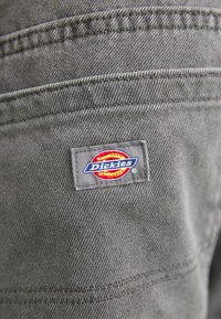 Gray fabric with a diagonal pattern features a square Dickies logo tag in red, yellow, and blue at the right side, near the seam.