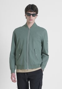 Zelena bomber jakna s prednjim zatvaračem, dva bočna džepa, rebrastim manžetama i dodatnim džepom na lijevoj rukavu.