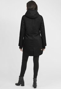 Manteau long noir avec capuche, tissu texturé, poignets à boutons et accent de label. Porté par-dessus un pantalon noir et des bottes à talons.