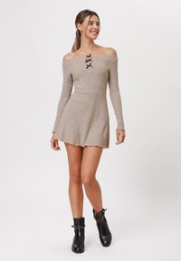 Beige off-the-shoulder ribbad miniklänning med svarta snörningsdetaljer, långa ärmar och en utsvängd kjol. Bärs med svarta ankelstövlar.