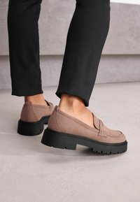 Mocassini in suede marrone con suola bassa e spessa nera, caratterizzati da un dettaglio cucito sulla parte anteriore. Indossati con pantaloni neri attillati su uno sfondo neutro.