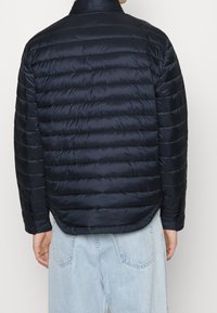 Mężczyzna w ciemnoniebieskiej pikowanej kurtce widzianej z tyłu, w połączeniu z jasnoniebieskimi dżinsami.