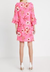 Nainen, joka on pukeutunut vaaleanpunaiseen kukalliseen polvipituiseen mekkoon, jossa on kerrokselliset kellohihat, ja hänellä on valkoiset korkokengät nilkkahihnalla. Hän on kääntynyt pois.