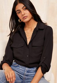 Black button-up shirt with two chest pockets, rolled sleeves, and a soft texture, paired with high-waisted light blue jeans.