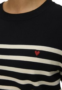 Maglione a righe bianco e nero con scollo rotondo, realizzato in materiale morbido, con un piccolo ricamo a forma di cuore rosso sul petto.