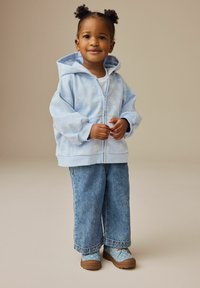 Lyseblå zip-up hoodie med blomstermønster, parret med brede denimbukser og mønstrede blå sko. Blødt stof, afslappet pasform.