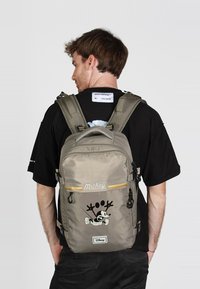 Mochila gris con un diseño impreso de Mickey Mouse, múltiples correas ajustables, textura suave y cremallera de acento amarillo.