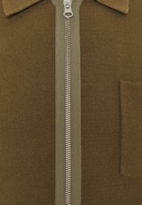 Vêtement en tissu brun côtelé avec une fermeture éclair et un col verts contrastants. Comprend une fermeture à glissière et une petite poche plaquée sur le côté gauche.