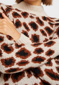 Strickpullover mit einem cremefarbenen Grund und großen braunen und schwarzen Leopardenflecken, geripptem Ausschnitt und Bündchen. Weiche, strukturierte Oberfläche.