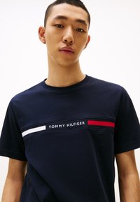 Junger Mann trägt ein dunkelblaues Tommy Hilfiger T-Shirt mit weißen und roten Streifen und Logo auf der Brust, schaut nach vorne.