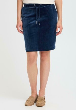 Jupe en velours côtelé bleu marine avec une coupe ajustée, taille à cordon de serrage et texture à côtes verticales. Associée à des mocassins beiges.
