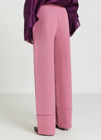 Weit geschnittene rosa Hosen mit umgeschlagenen Säumen, getragen mit einer dunkelvioletten Bluse und schwarzen spitzen Schuhen, von hinten gezeigt.