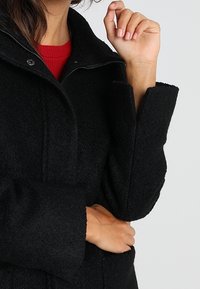 Svart ullrock med hög krage, tryckknappsöppningar och vida ärmar, med en mjuk textur och subtila sömnadsdetaljer.