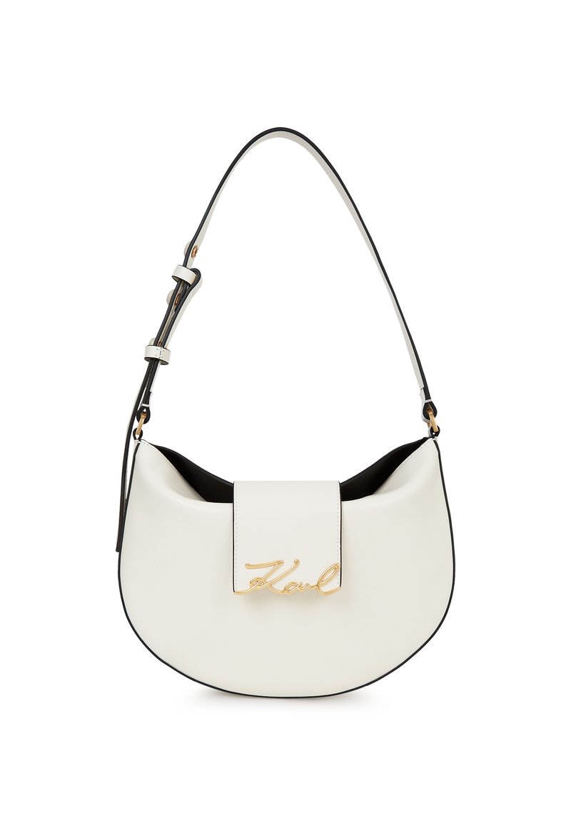 Balta pusmēness formas soma ar regulējamu siksniņu un zelta burtiem "Karl" logotipu priekšējā atloks uz vienkārša balta fona.