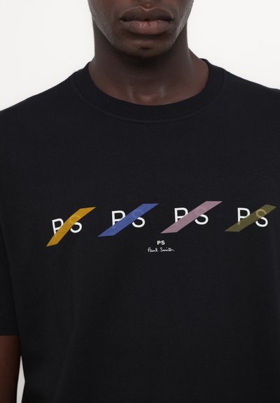 Črna bombažna majica s kratkimi rokavi z ponovljenim logotipom "PS" v rumenih, modrih, roza in zelenih diagonalnih črtah. Klasičen ovratnik s krožnim izrezom.