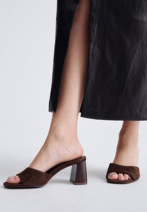 Pies descalzos con tacones mule de ante marrón abiertos en la puntera, combinados con pantalones negros de pierna ancha con aberturas laterales.