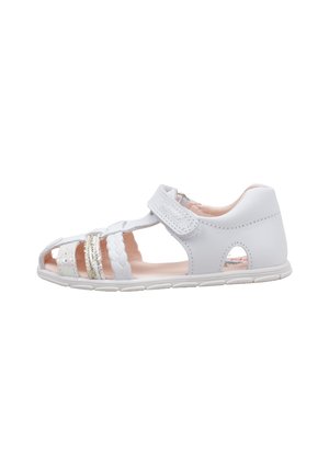Sandalia blanca para niño pequeño con tiras trenzadas y metálicas, cierre de velcro en el tobillo y diseño de punta abierta sobre una suela de goma blanca.