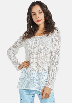 Suéter blanco de encaje con patrones florales, mangas largas y escote amplio, combinado con pantalones de patrón azul claro.