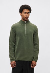 Πράσινο πλεκτό πουλόβερ με ψηλό γιακά και κλείσιμο με μισό φερμουάρ. Διαθέτει αντίθετη ραφή κατά μήκος των μανικιών. Μαλακή υφή, άνετη εφαρμογή.