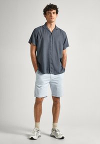 Camicia a maniche corte, grigia scuro, abbottonata, abbinata a pantaloni corti azzurri chiari. Il modello indossa sneaker bianche e calzini crema, con le mani nelle tasche.