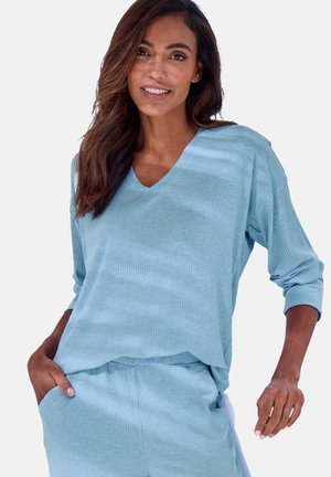 Woman with long hair wearing a light blue ribbed V-neck top and matching pants with hands in pockets, smiling gently.