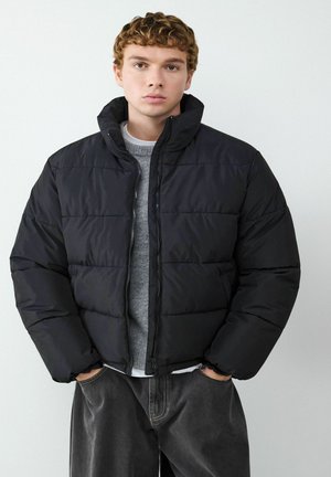 Schwarze Steppjacke mit hohem Kragen und Reißverschluss, mit horizontaler Naht und lässiger Passform, getragen über einem grauen Pullover.