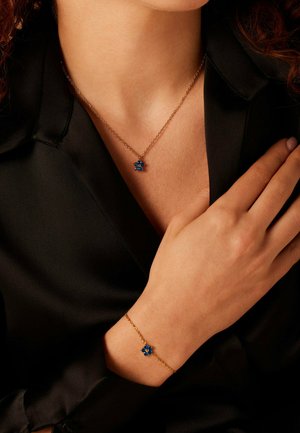 Gouden ketting en armband met een blauwe edelsteen in de vorm van een bloem als hanger. De textuur is glad met een verfijnd ontwerp en fijne schakels.