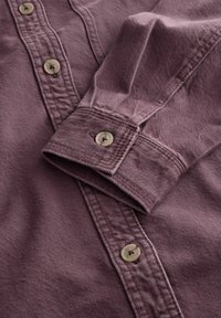 Gros plan sur un poignet de chemise à manches longues plié et une patte de boutonnage en tissu texturé violet atténué avec des boutons beige.