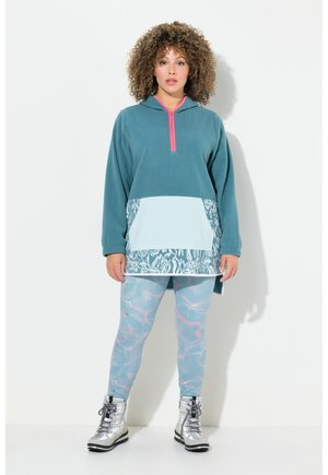 Tüm grüner Fleece-Pullover mit pinkem Reißverschluss und weißem gemustertem Saum, kombiniert mit hellblauen, marmorierten Leggings und silbernen Knöchelschuhen.