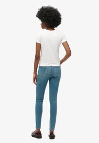 Camiseta blanca de manga corta, diseño ajustado, combinada con jeans ajustados azules y zapatos marrones, vista desde atrás. Apariencia limpia y minimalista.