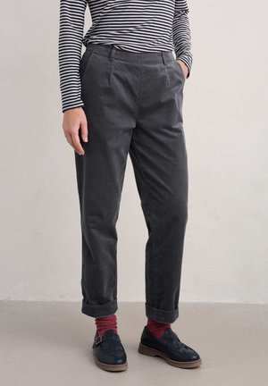 Osoba ubrana w podwinięte ciemnoszare spodnie, granatowe buty na klamrę, czerwone skarpetki oraz czarno-biały pasiasty top z długim rękawem, stojąca na jasnej podłodze.