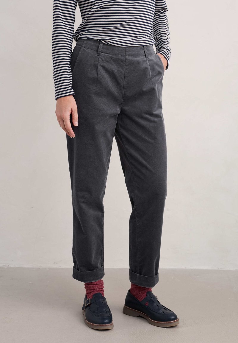 Personne portant un pantalon gris foncé retroussé, des chaussures bleu marine à boucle, des chaussettes rouges et un haut rayé noir et blanc à manches longues, debout sur un sol clair.