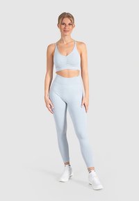 Brassière de sport bleu clair avec détail froncé, associée à des leggings taille haute assortis. Les deux sont fabriqués en tissu extensible, portés avec des baskets blanches.