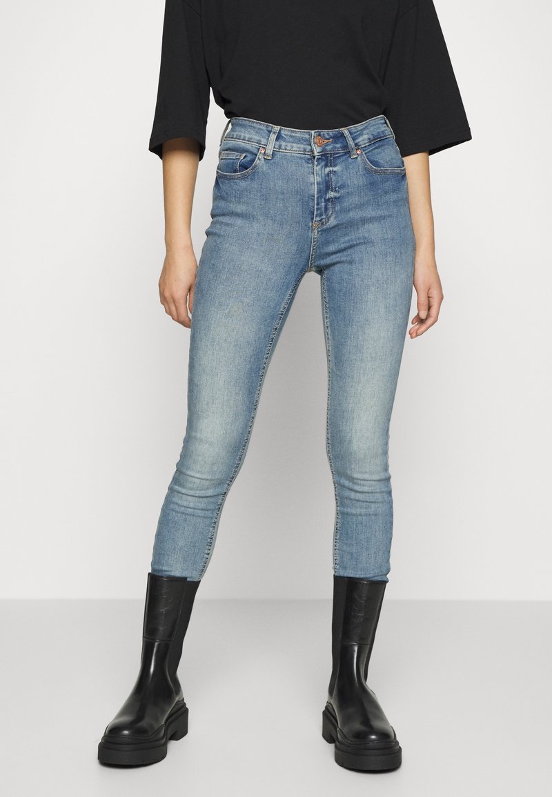 Personne portant un jean skinny bleu clair, des bottes noires épaisses à mi-mollet et un haut noir à manches longues, debout devant un fond uni.