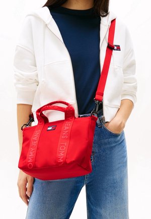 Borsa a mano rossa in nylon con doppi manici e tracolla staccabile. Presenta la scritta in rilievo "TOMMY JEANS" sulle cinghie e una patch con il logo.