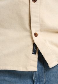 Cămașă din corduroy bej cu nasturi maro, având o textură de dungi verticale și un etichetă pe lateral; asortată cu blugi albaștri.